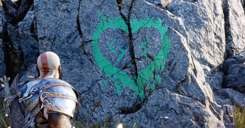 A screenshot of the game God of War Ragnarok, where the player looks at a green heart symbol painted on the side of a rock, containing Nordic symbols that translate to "SH" and "JS"