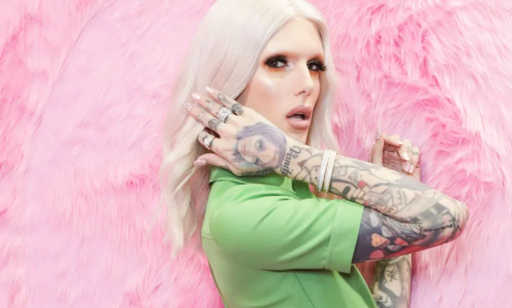 Jeffree Star wearing a green suit and sporting long blonde hair standing against a pink background.
