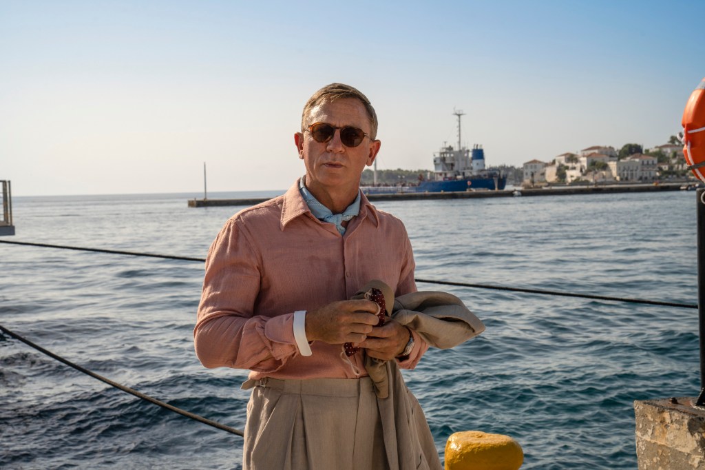 A still from the movie Glass Onion: A Knives Out Mystery shows actor Daniel Craig playing detective Benoit Blanc wearing a pink shirt, light brown trousers and sunglasses as he stands on a boat