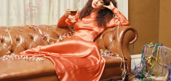 Kate Bush wearing an orange dress and holding a glass of wine while lounging on a sofa in 1979 for her BBC Christmas special.