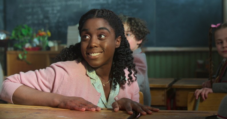 A still from Netflix's Matilda: The Musical showing actor Lashana Lynch as Miss Honey wearing a turquoise shirt and pink cardigan sitting a desk in a classroom