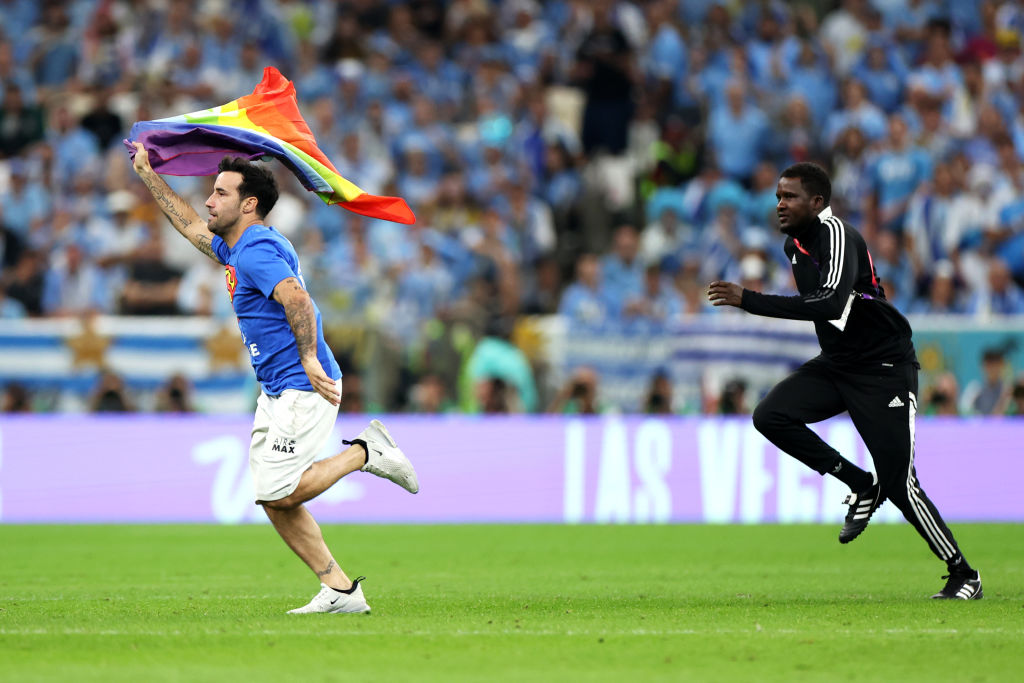 A photo of a pitch invader wearing a shirt reading "Save Ukraine" as he holds a rainbow flag during the FIFA World Cup Qatar 2022.