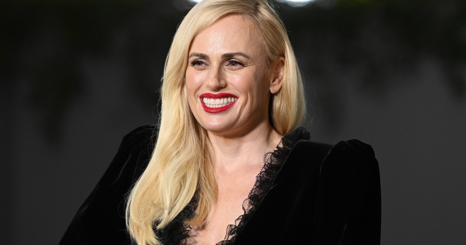 Photo of actor Rebel Wilson dressed in a black dress smiles for the cameras at a press event