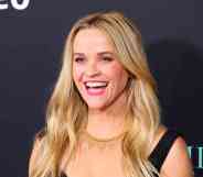 A headshot of actor Reese Witherspoon wearing a black top and smiling as she has her photo taken at a red carpet event