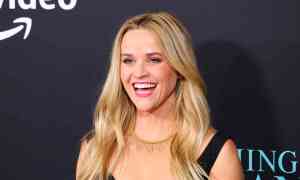 A headshot of actor Reese Witherspoon wearing a black top and smiling as she has her photo taken at a red carpet event