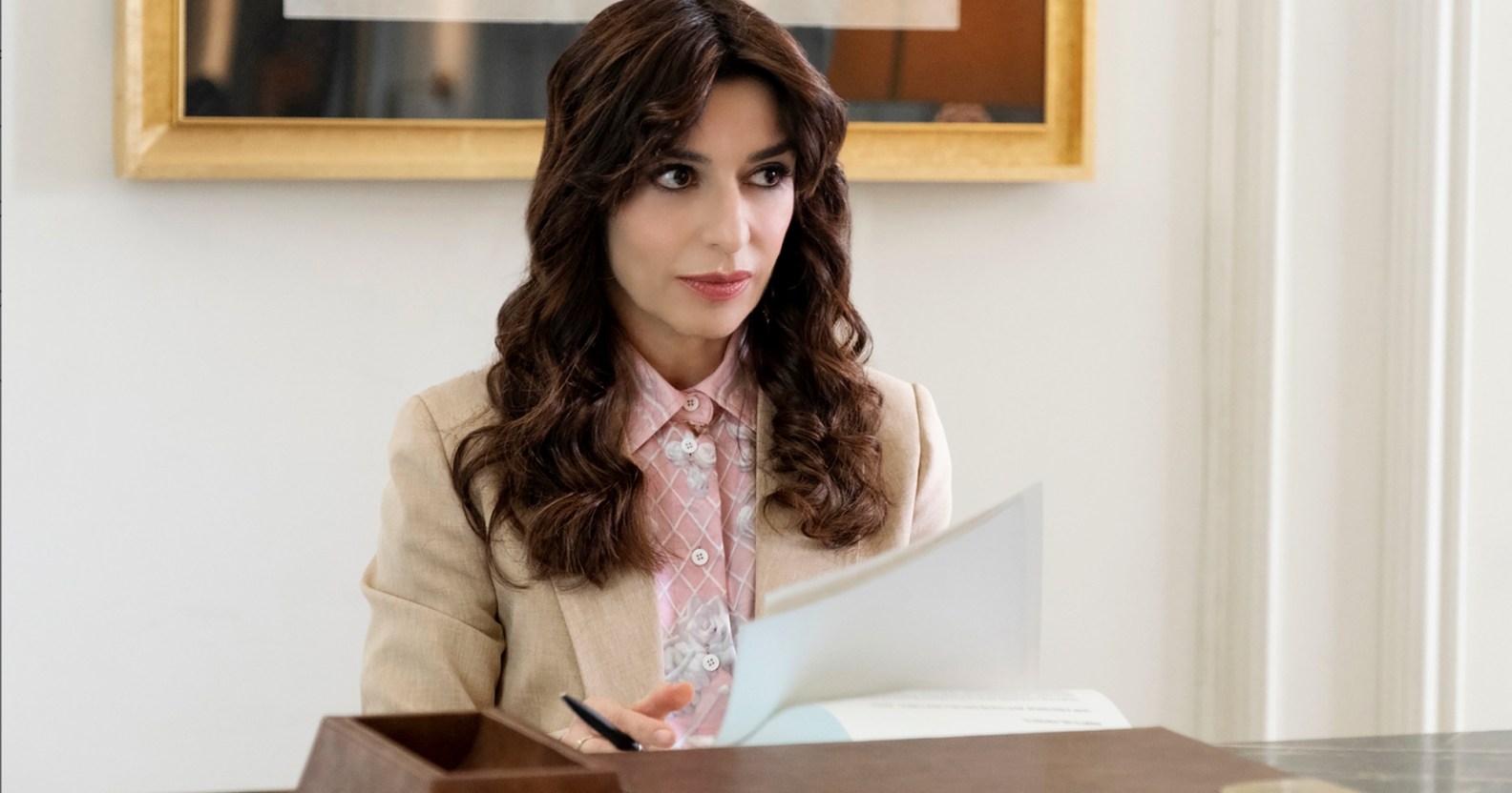 A still from HBO's The White Lotus shows actor Sabrina Impacciatore as Valentina dressed in a cream suit jacket and pink shirt standing behind a reception desk holding some papers.