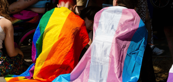 Two people sitting side by side, one in a rainbow flag and one in a trans flag