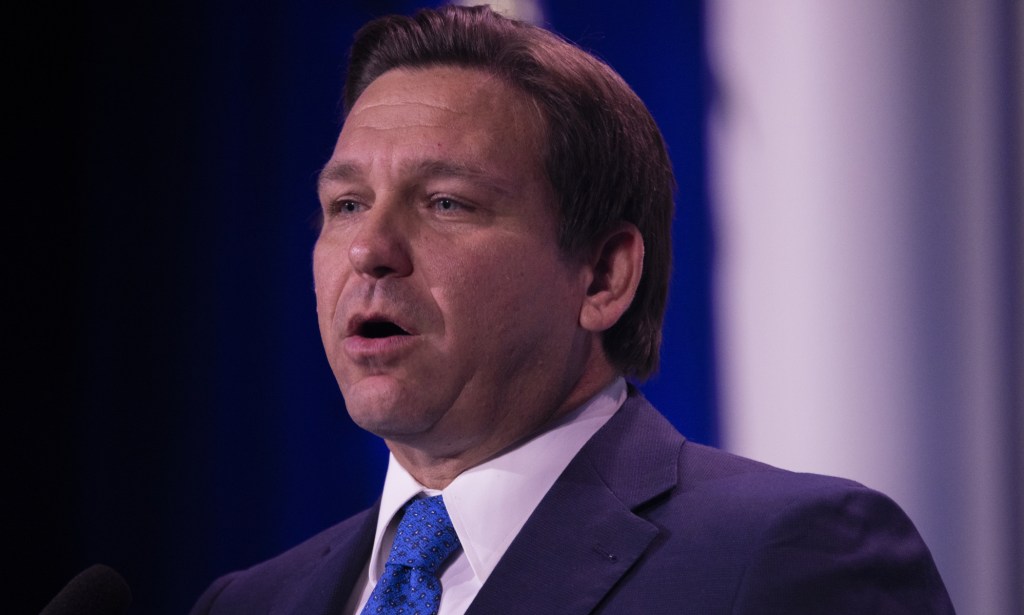Ron DeSantis speaks to a crowd while wearing a black suit and blue tie.