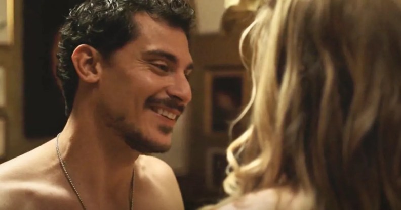 A still from The White Lotus showing actor Stefano Gianino as Niccolo smiling as he strips down in front of a woman with blonde hair who is turned away from the camera