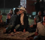 Still from TV series Yellowstone showing actor Lilli Kay as Clara wearing a denim jacket and jeans kissing a woman wearing a black coat, white shirt and white cowboy hat