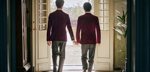 An image taken from Netflix's Instagram shows Young Royals actors Edvin Ryding and Omar Rudberg as Crown Prince Wilhelm and Simon dressed in their boarding school uniforms and walking hand in hand through some French patio doors with their backs to the camera
