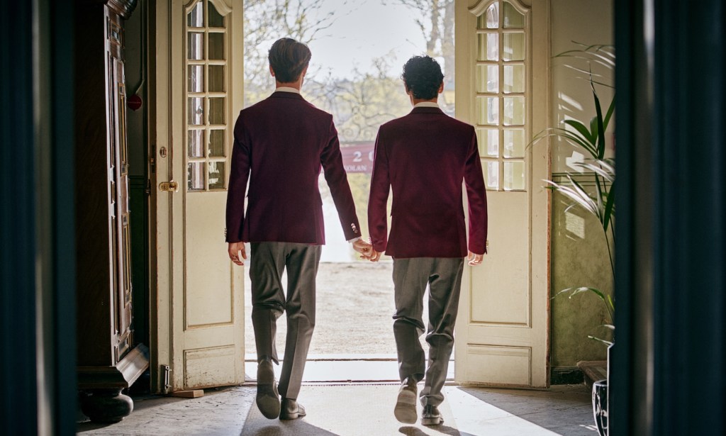 An image taken from Netflix's Instagram shows Young Royals actors Edvin Ryding and Omar Rudberg as Crown Prince Wilhelm and Simon dressed in their boarding school uniforms and walking hand in hand through some French patio doors with their backs to the camera