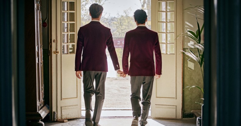 An image taken from Netflix's Instagram shows Young Royals actors Edvin Ryding and Omar Rudberg as Crown Prince Wilhelm and Simon dressed in their boarding school uniforms and walking hand in hand through some French patio doors with their backs to the camera