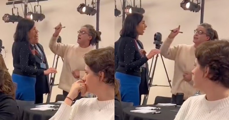A woman screams and points at the back of a room of people sitting at tables, who appear very uncomfortable