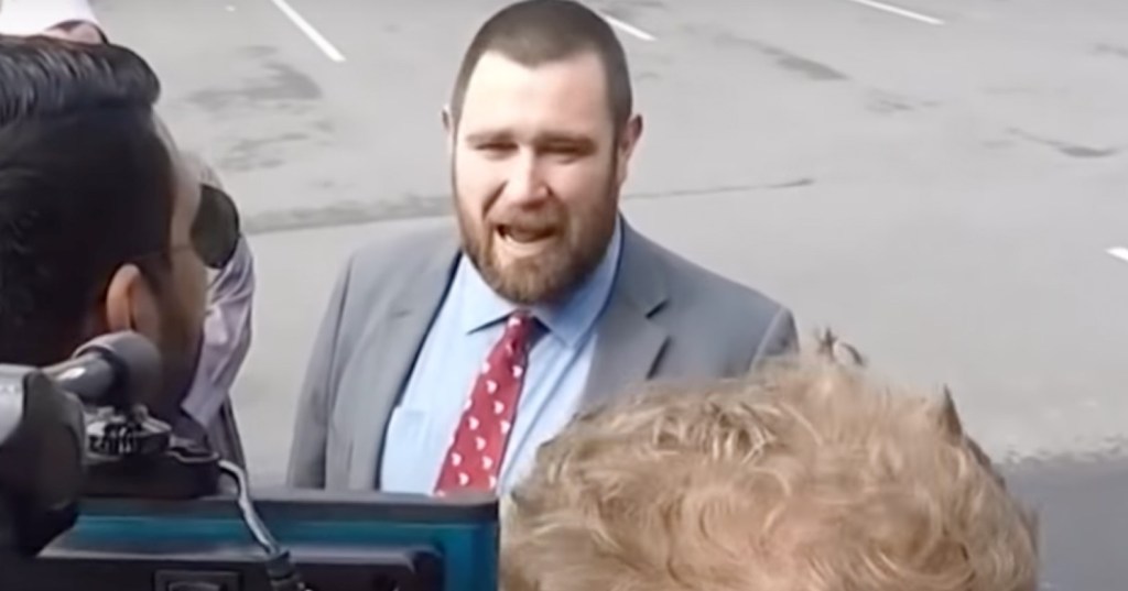 New Zealand pastor Logan Robertson, wearing a grey suit jacket over a light blue shirt with red patterned tie, speaks to cameras in a parking lot