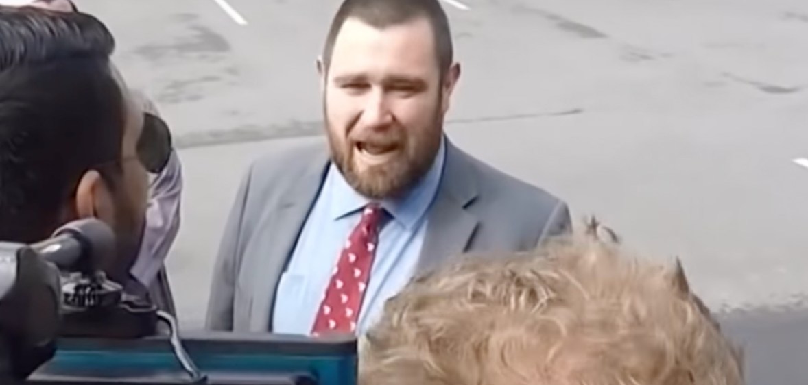 New Zealand pastor Logan Robertson, wearing a grey suit jacket over a light blue shirt with red patterned tie, speaks to cameras in a parking lot