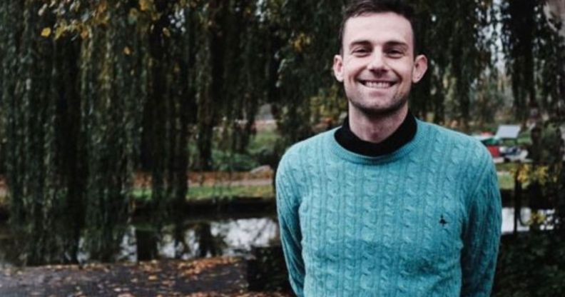 Photo of Christian teacher Joshua Sutcliffe wearing a turquoise top smiling as he stands in a countryside setting