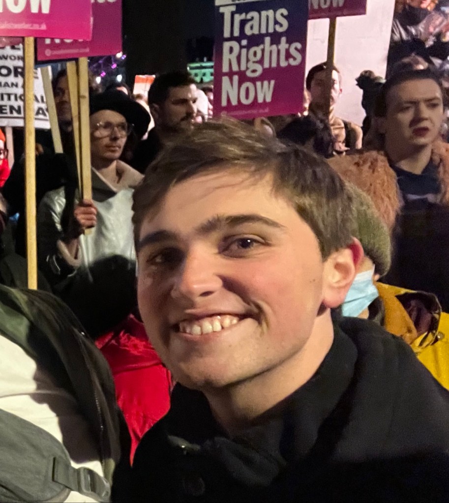 Alex Charilaou at a trans rights protest. They are pictured with people in the background holding placards that read "trans rights now".
