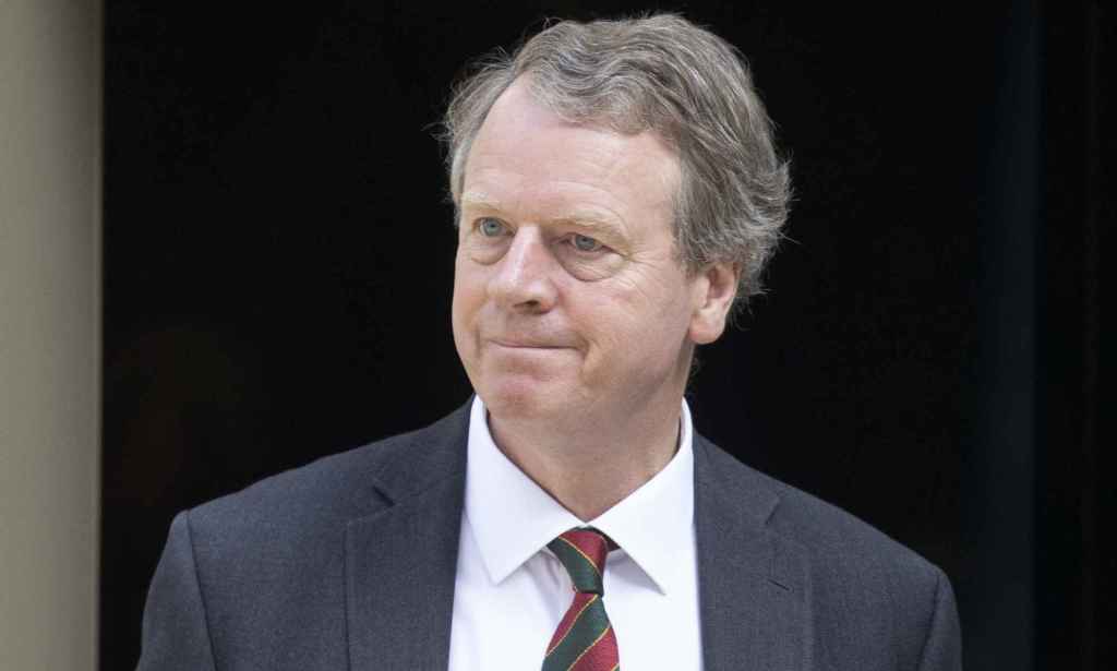 Alister Jack in a grey suit, white shirt and red tie