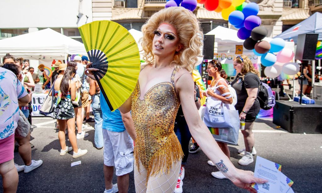 A drag queen uses a yellow hand fan during a crowded Pride event