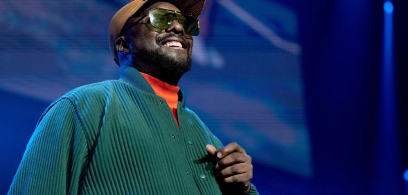 will.i.am, wearing a green sweater and brown brim cap, smiles to an audience while on stage at a concert.