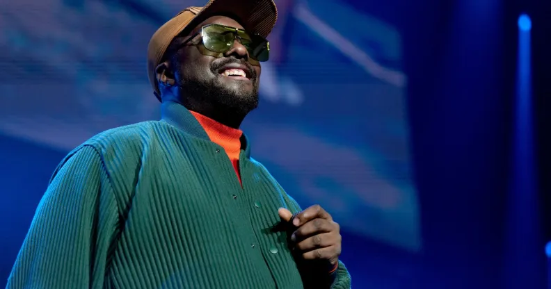 will.i.am, wearing a green sweater and brown brim cap, smiles to an audience while on stage at a concert.