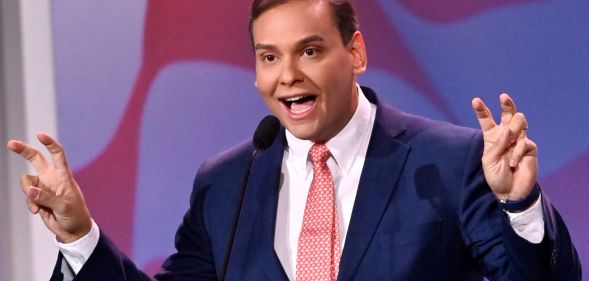 Photo of US congressman George Santos wearing a navy suit, white shirt and pink tie as he makes air quotation gestures with both of his hands.