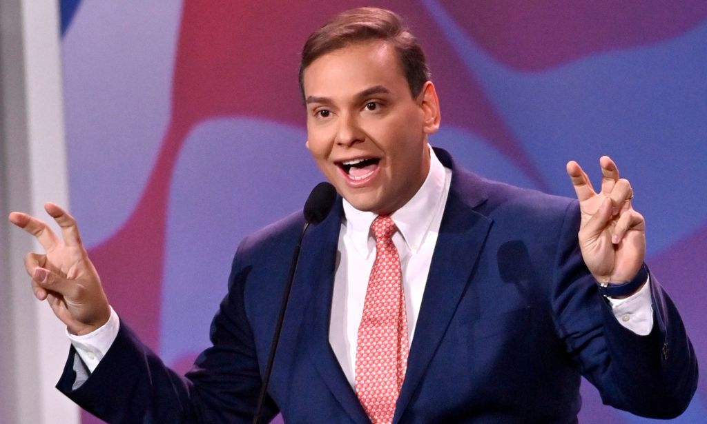 Photo of US congressman George Santos wearing a navy suit, white shirt and pink tie as he makes air quotation gestures with both of his hands.