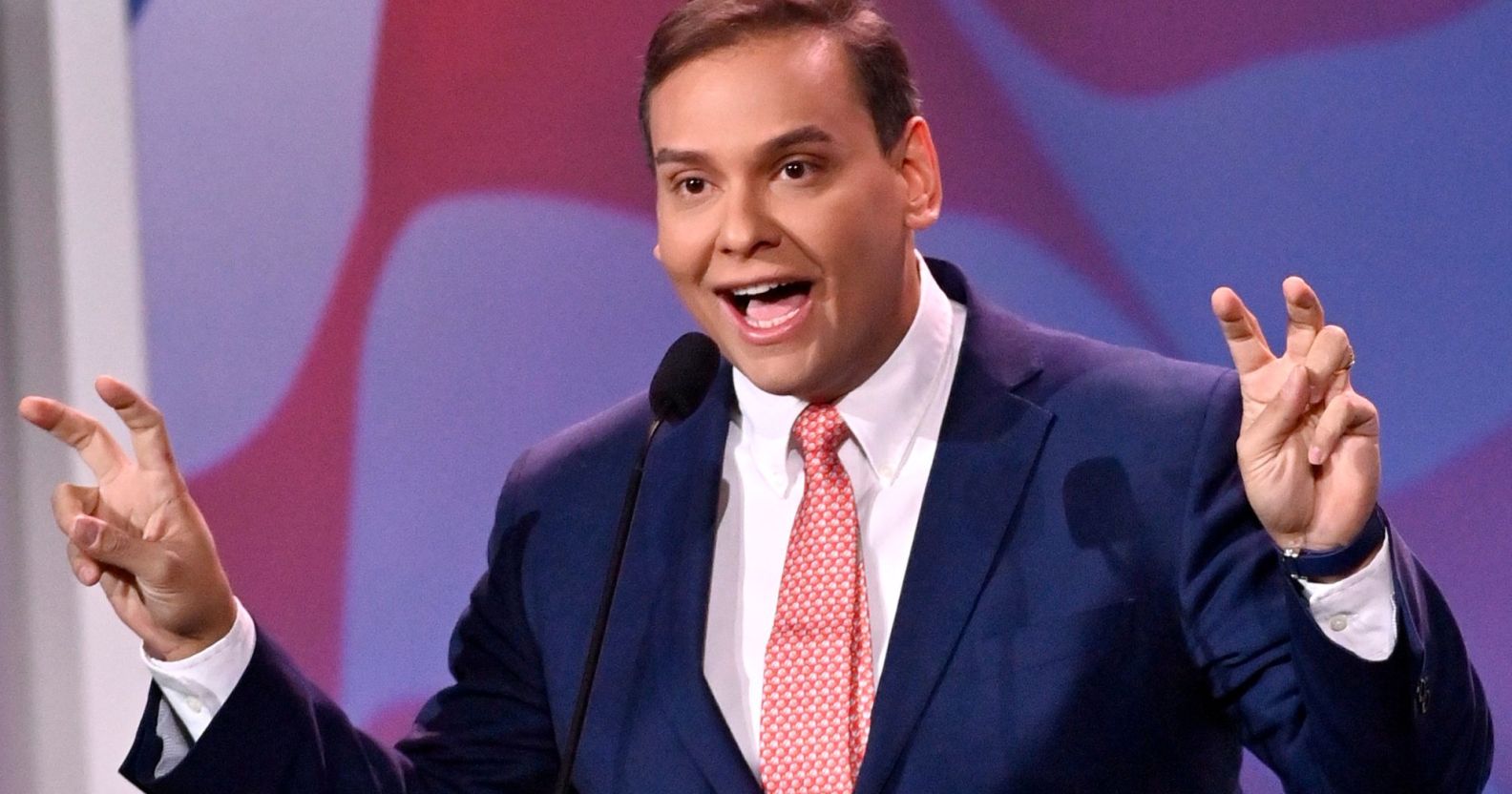Photo of US congressman George Santos wearing a navy suit, white shirt and pink tie as he makes air quotation gestures with both of his hands.
