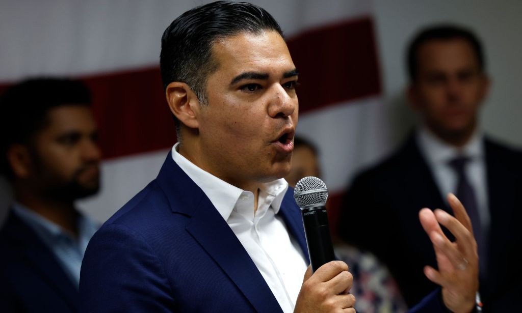 Robert Garcia, holding a microphone, speaks to a group of people in an open collar blue blazer.
