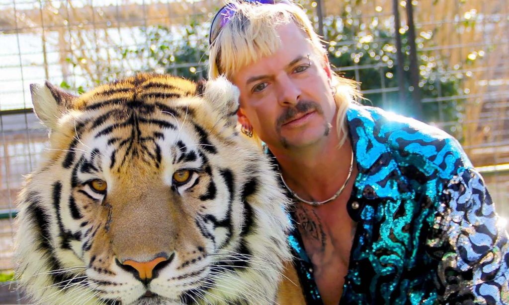 Joe Exotic hugs a tiger while wearing a blue and white shiny jacket.