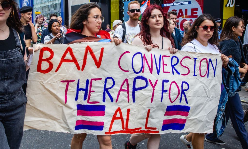A photo shows LGBTQ+ activists holding a sign reading "ban conversion therapy for all."