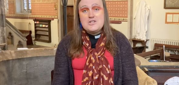 Bingo Allison, a non-binary, genderqueer Church of England priest, wears a red top, sweater and scarf while they speak about religion and Trans Day of Visibility