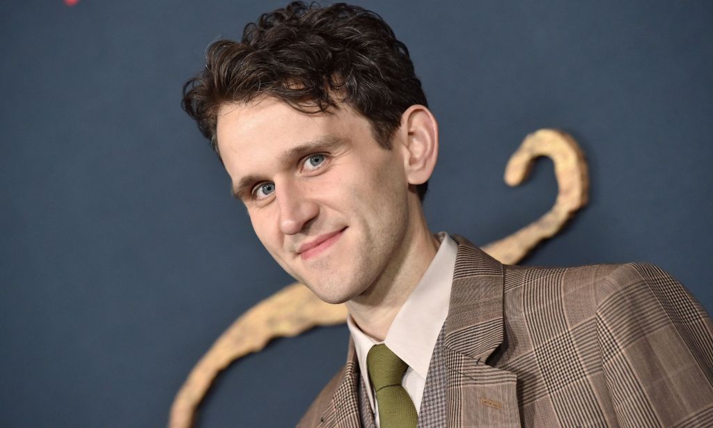 Actor Harry Melling, who rose to fame as Dudley Dursley in the Harry Potter franchise, wears a brown patterned suit jacket with a green-brown tie as he smiles at the camera
