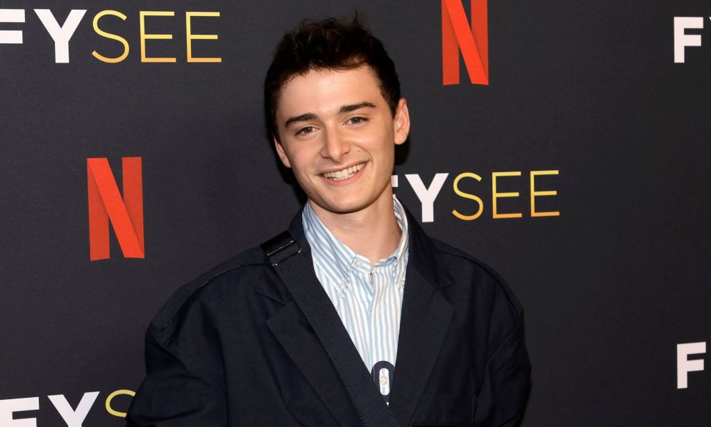 Noah Schnapp smiles while wearing a light coloured shirt and dark coloured jacket while standing in front of a background with the Netflix logo on it