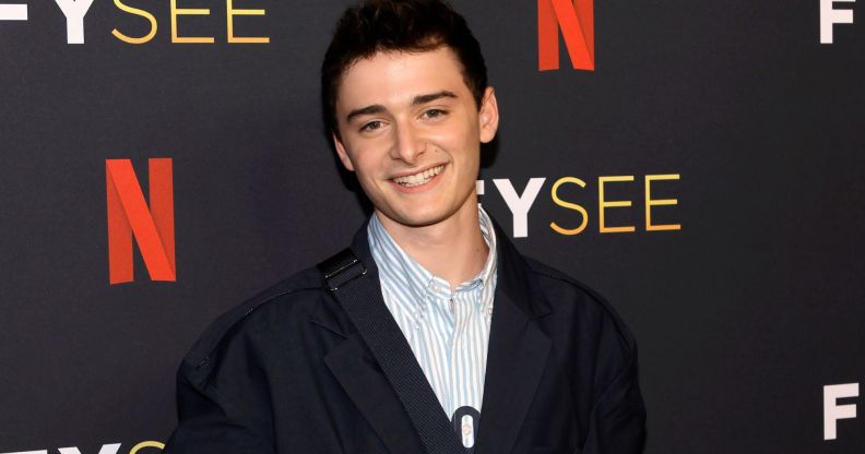 Noah Schnapp smiles while wearing a light coloured shirt and dark coloured jacket while standing in front of a background with the Netflix logo on it