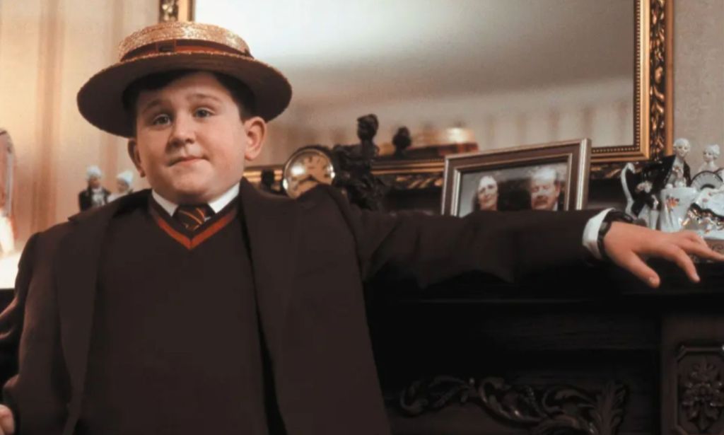 Harry Melling wears a red British school uniform complete with a straw cap and resting his arm against a fireplace while portraying Dudley Dursley in the Harry Potter franchise