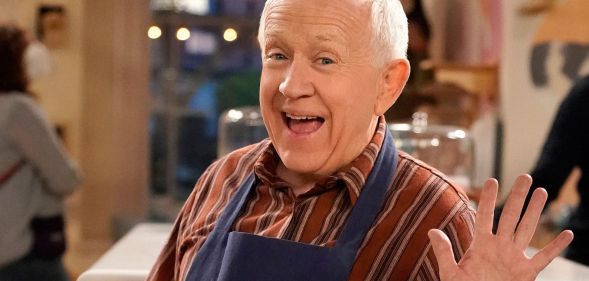 Leslie Jordan smiles and waves while wearing a red striped shirt and baking apron on the set of Call Me Kat