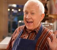 Leslie Jordan smiles and waves while wearing a red striped shirt and baking apron on the set of Call Me Kat