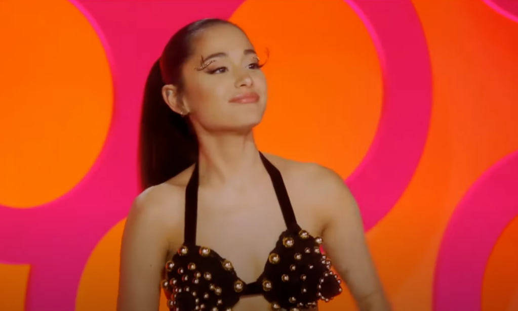 Ariana Grande wears a black bikini like top with copper pears decorating it as she sits on the Drag Race season 15 judges panel