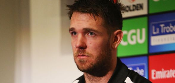 A photo shows Former Australian Football League (AFL) star Dane Swan wearing a black and white top sitting in a room with the names of football sponsors on the wall behind him
