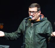 A photo shows singer-songwriter Paul Heaton wearing a green coat and standing behind a microphone on stage as he holds his hands up