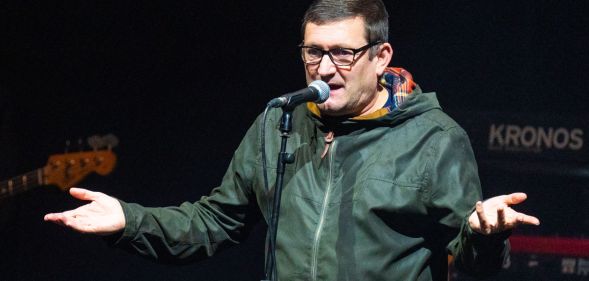 A photo shows singer-songwriter Paul Heaton wearing a green coat and standing behind a microphone on stage as he holds his hands up
