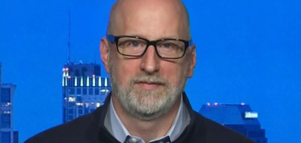 A close-up photo of attorney David French wearing a black top over a blue shirt sitting by a window which has skyscrapers in the background
