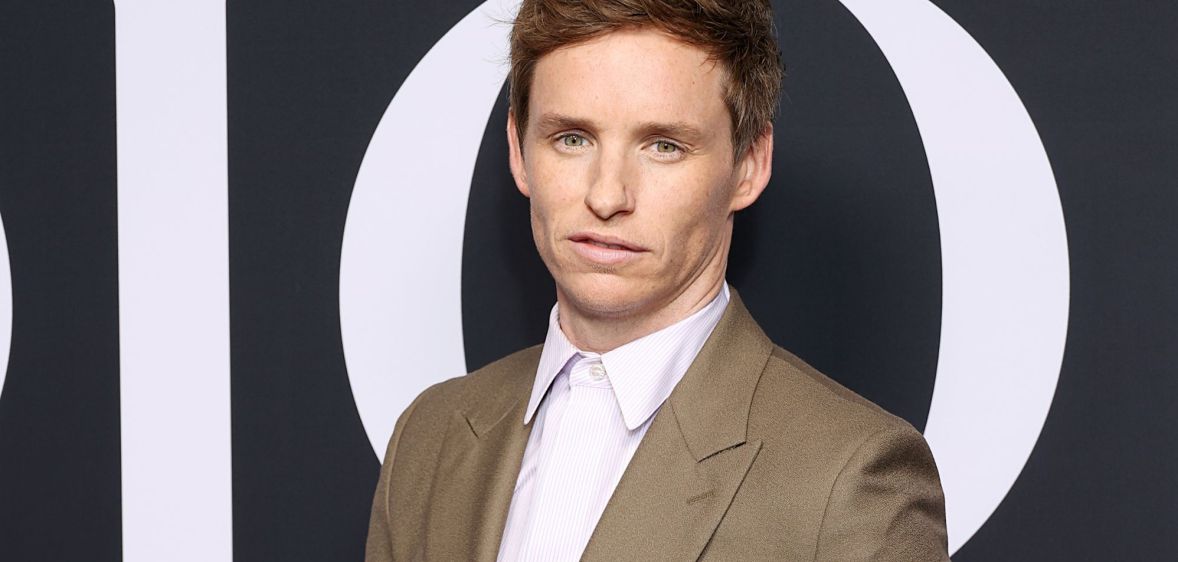 Eddie Redmaybe wears a white button-up shirt with a light brown jacket as he poses for the camera at an event