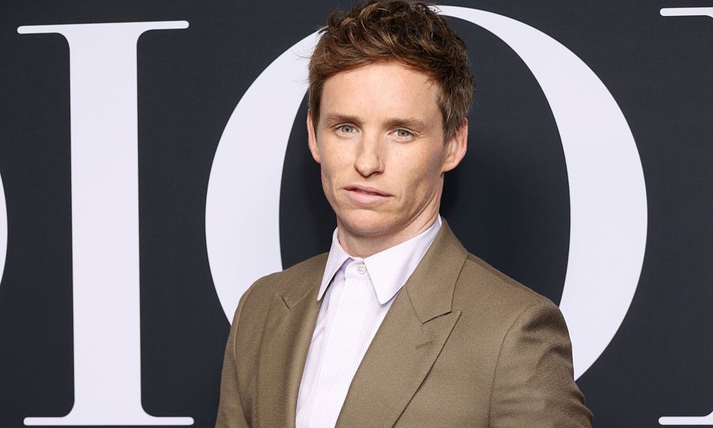 Eddie Redmaybe wears a white button-up shirt with a light brown jacket as he poses for the camera at an event