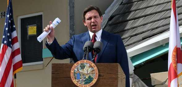 A photo of Florida governor Ron DeSantis wearing a navy suit, white shirt and red tie speaking at a podium