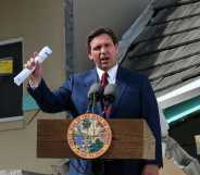 A photo of Florida governor Ron DeSantis wearing a navy suit, white shirt and red tie speaking at a podium