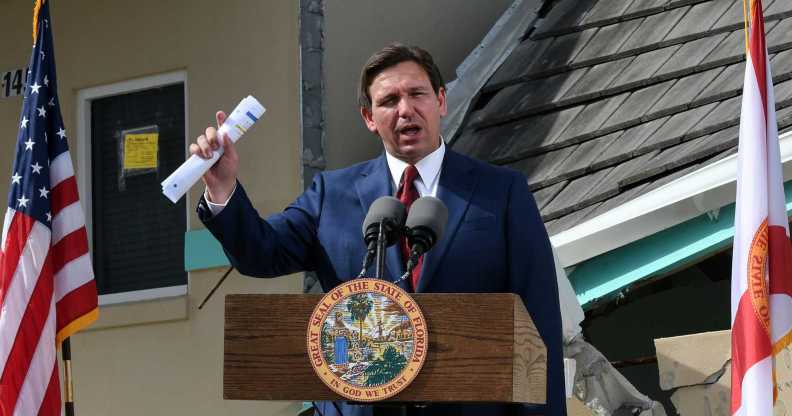 A photo of Florida governor Ron DeSantis wearing a navy suit, white shirt and red tie speaking at a podium