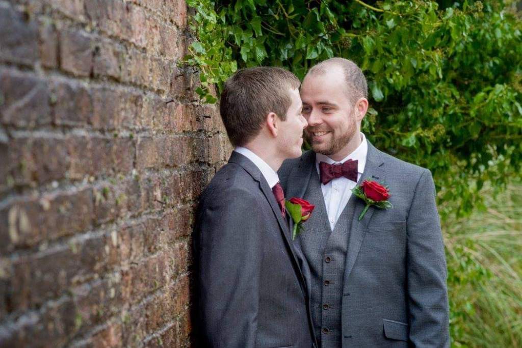 Conversion therapy survivor Garry Adair-Gilliland is pictured on the right hand side leaning close to his husband on their wedding day. His husband is leaning against a red brick wall which is visible on the left hand side of the picture. In the background is shrubbery and greenery.